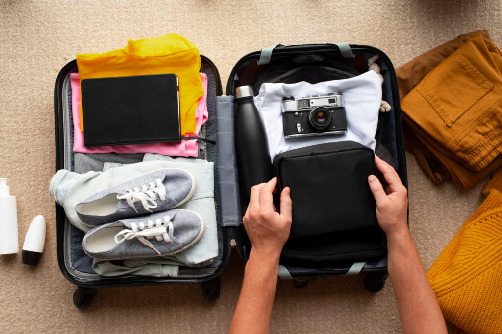 Packing road trip travel tips with a top view of an open suitcase being packed with travel essentials including clothes, shoes, a camera, and a toiletry bag on a carpeted floor.