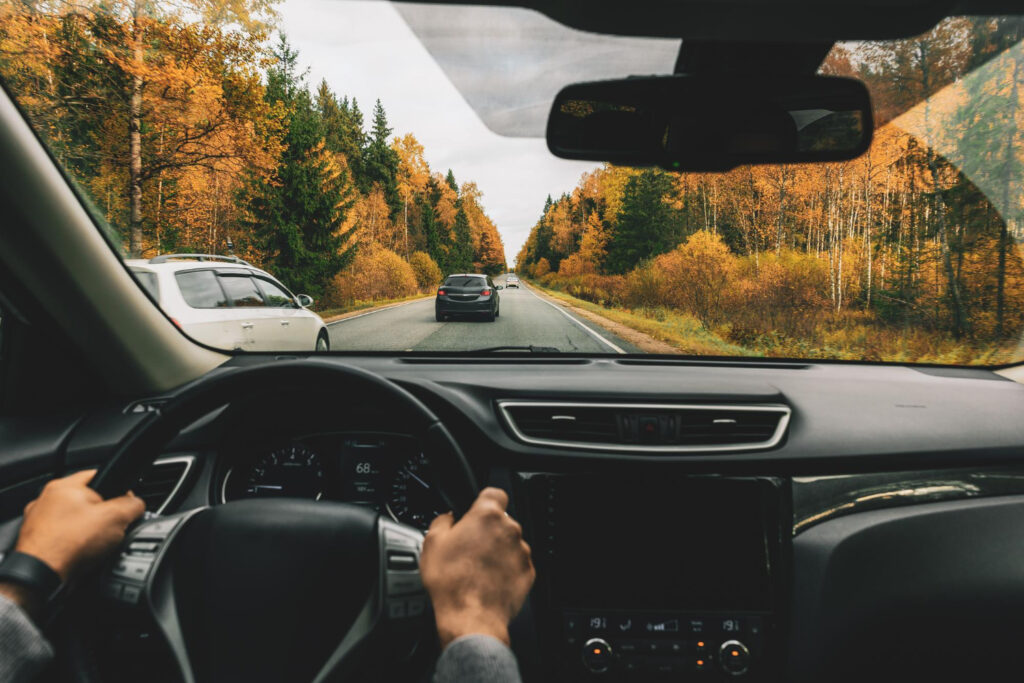 A driver's perspective from inside a car driving on a forest road surrounded by autumn foliage. The dashboard shows a modern vehicle interior with a clear view of the road ahead, where other cars are visible traveling in the same direction. The trees alongside the road display a vivid array of fall colors, with bright yellows, oranges, and greens dominating the scene. This image captures the essence of a road trip during the autumn season, showcasing the beauty of driving through a wooded area with seasonal foliage.