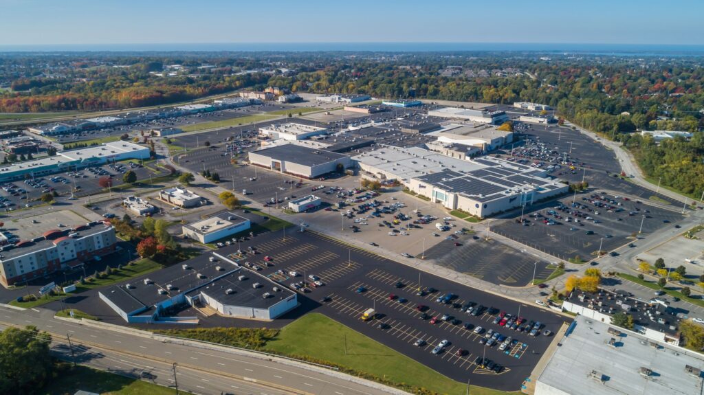 Shopping erie pennsylvania with an aerial view of a sprawling commercial area with large parking lots and multiple buildings, including a prominent shopping mall with a flat roof featuring solar panels. The scene captures a mix of retail and hospitality facilities amidst an autumn landscape, with colorful trees marking the change of seasons. The image provides a comprehensive look at suburban commercial development and renewable energy use in retail architecture."