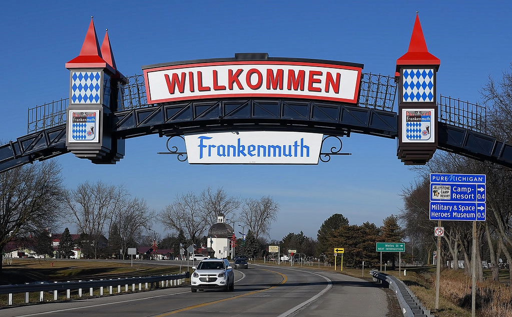 An image displaying a street view with a prominent welcome sign reading "WILLKOMMEN" in bold red letters, mounted on an overhead archway bridge. Below, the word "Frankenmuth" is featured in a decorative script, reinforcing the town's warm welcome. Clear blue skies provide the perfect backdrop for this inviting entrance, while vehicles are seen approaching on the road, symbolizing the steady flow of visitors to this beloved destination.