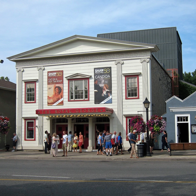 The image features the façade of The Royal George Theatre, a prominent venue for the Shaw Festival in Niagara-on-the-Lake, Ontario, Canada. The theater's classic architecture includes a white exterior with red accents and grandiose columns. A large banner above the entrance announces the 50th season of the Shaw Festival, with posters for the plays "Cat on a Hot Tin Roof" and "Candida." A group of patrons gathers in front of the red-painted doors, some engaged in conversation, while others are in line to enter. The scene is vibrant with the charm of a sunny day, and the sidewalk is adorned with lush hanging flower baskets, adding to the quaint and welcoming atmosphere of this historic cultural landmark.