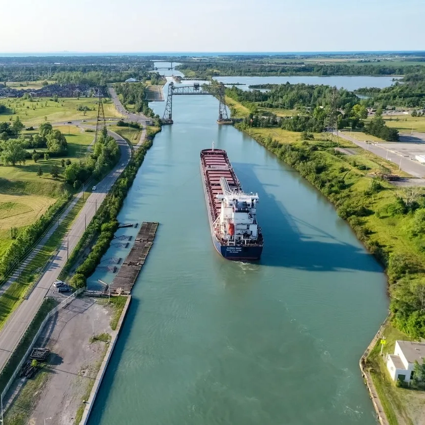 An aerial view of the Welland Canal with a large red-hulled cargo ship navigating through the waterway. The canal stretches straight through the image, bordered by green grassy areas on both sides and a road running parallel to its left. A series of distant bridges spans the canal, and the landscape is dotted with sparse buildings. The sky is clear and blue, suggesting a bright sunny day on a weekend in niagara falls. The water is calm and reflects a slight shimmer from the overhead sun.